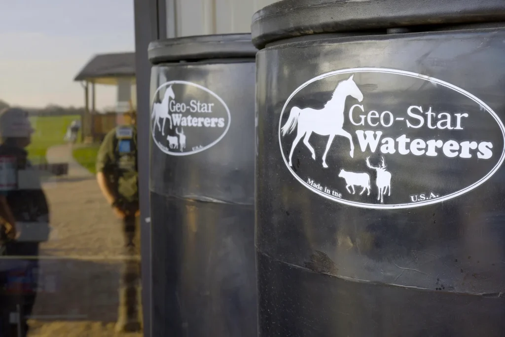 Close-up of black Geo-Star Waterers for livestock, featuring the brand logo with horse, cattle, and deer silhouettes, on display outdoors at Dutchman’s Farm Supply.