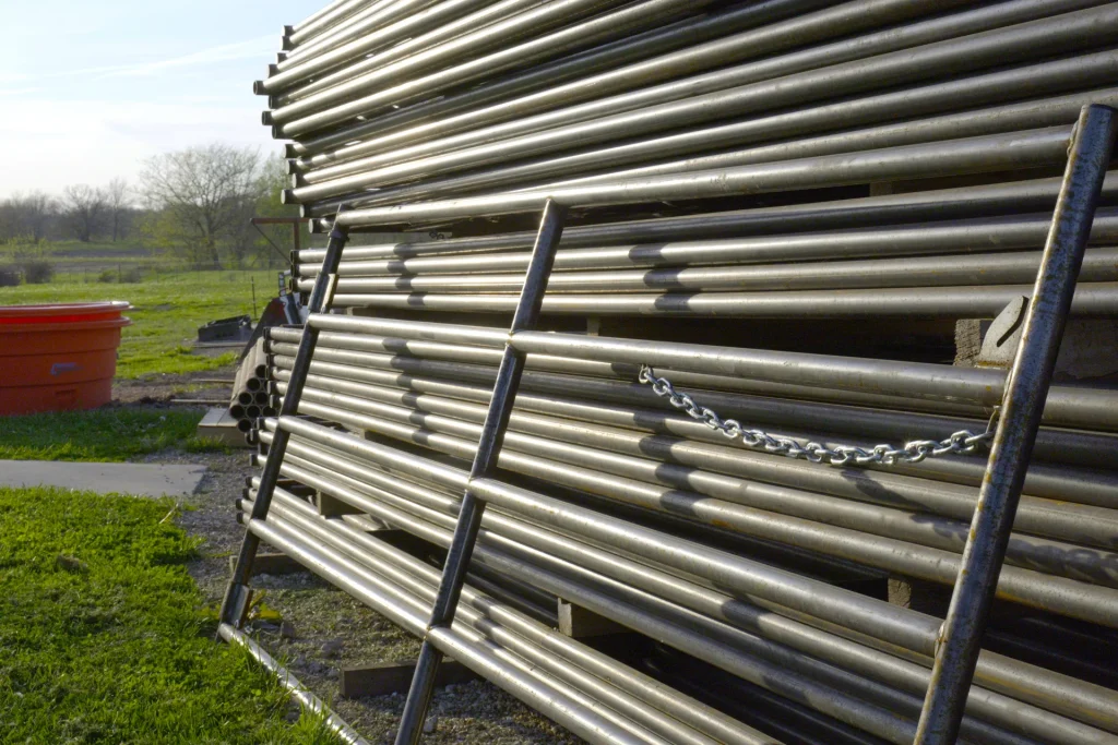 Large stacks of heavy-duty galvanized metal corral gates and steel fencing pipes stored outdoors on a gravel lot at Dutchman’s Farm Supply.