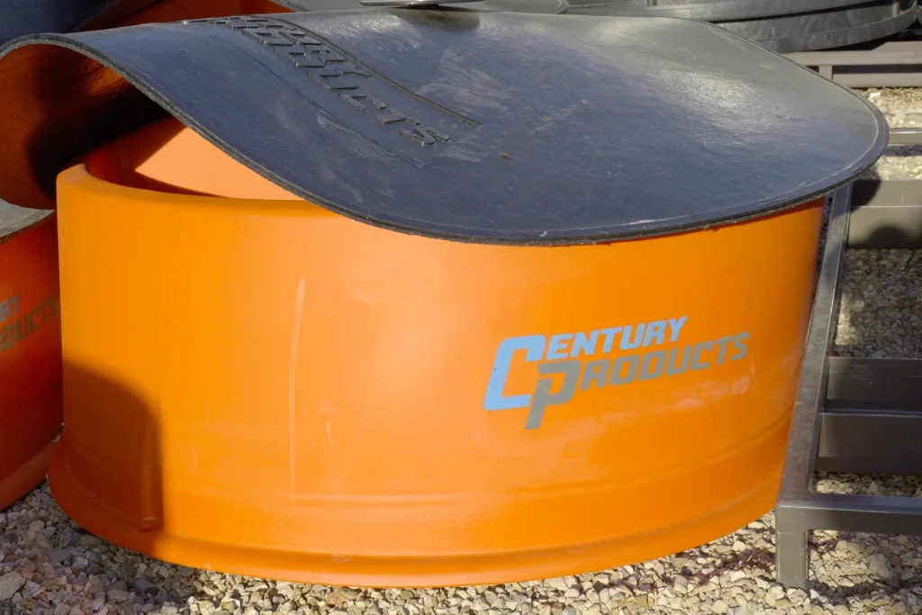 An orange Century Products mineral feeder with a heavy-duty black rubber lid, sitting on a gravel lot at Dutchman's Farm Supply.